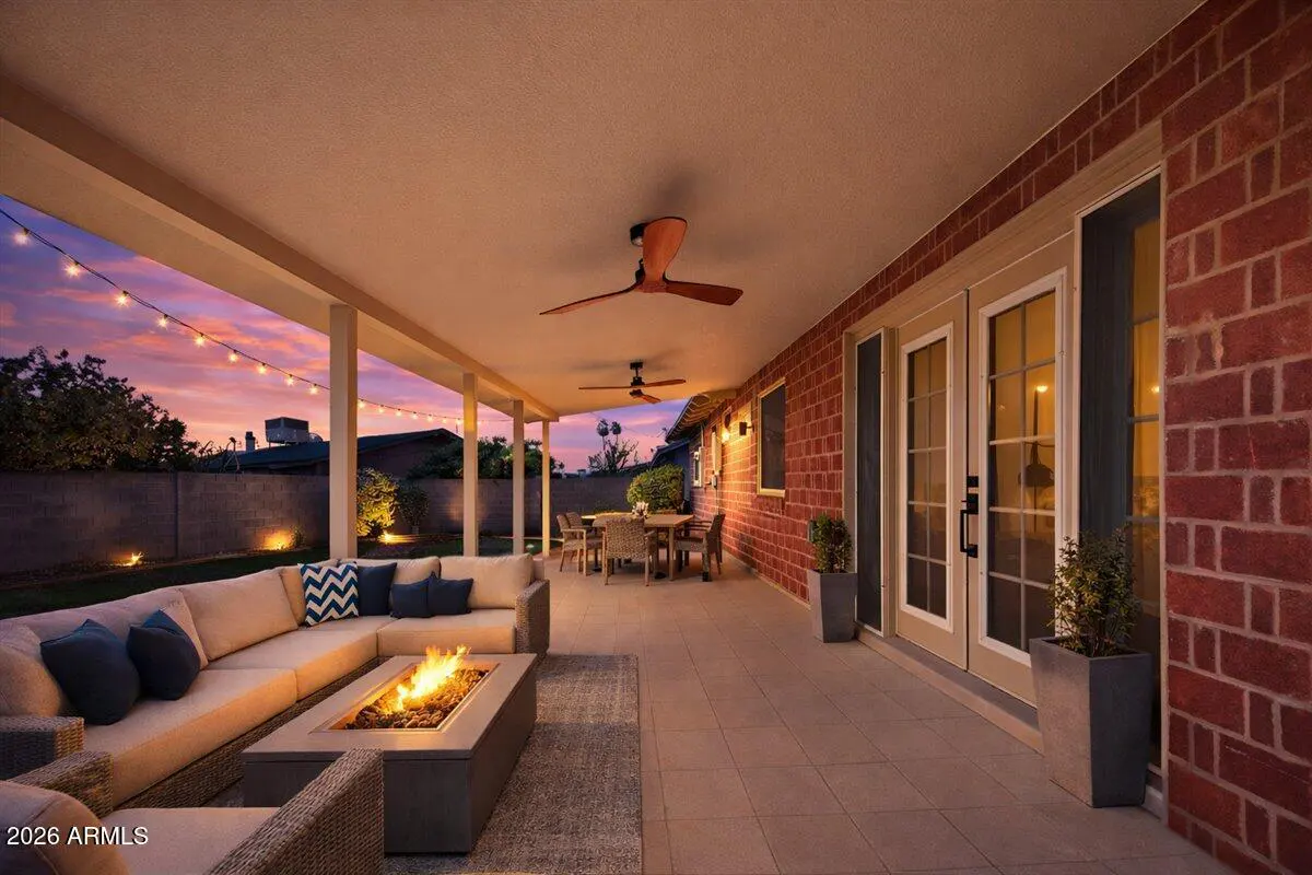 Covered patio with brick wall, ceiling fans, and string lights. A fire pit sits in front of a beige sectional sofa.