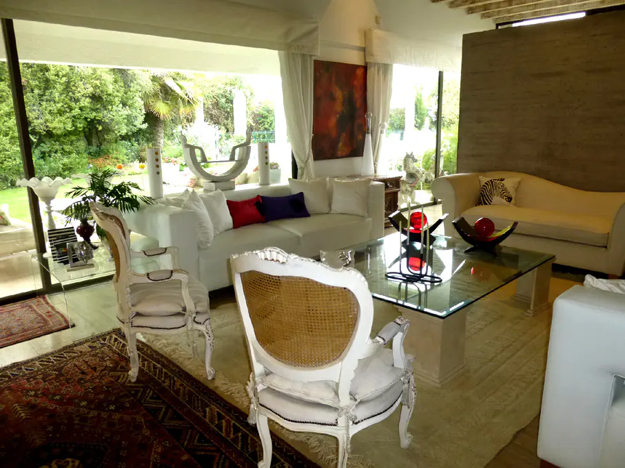 Bright living room with white sofas, glass table, and ornate chairs. Large windows overlook a lush green garden.