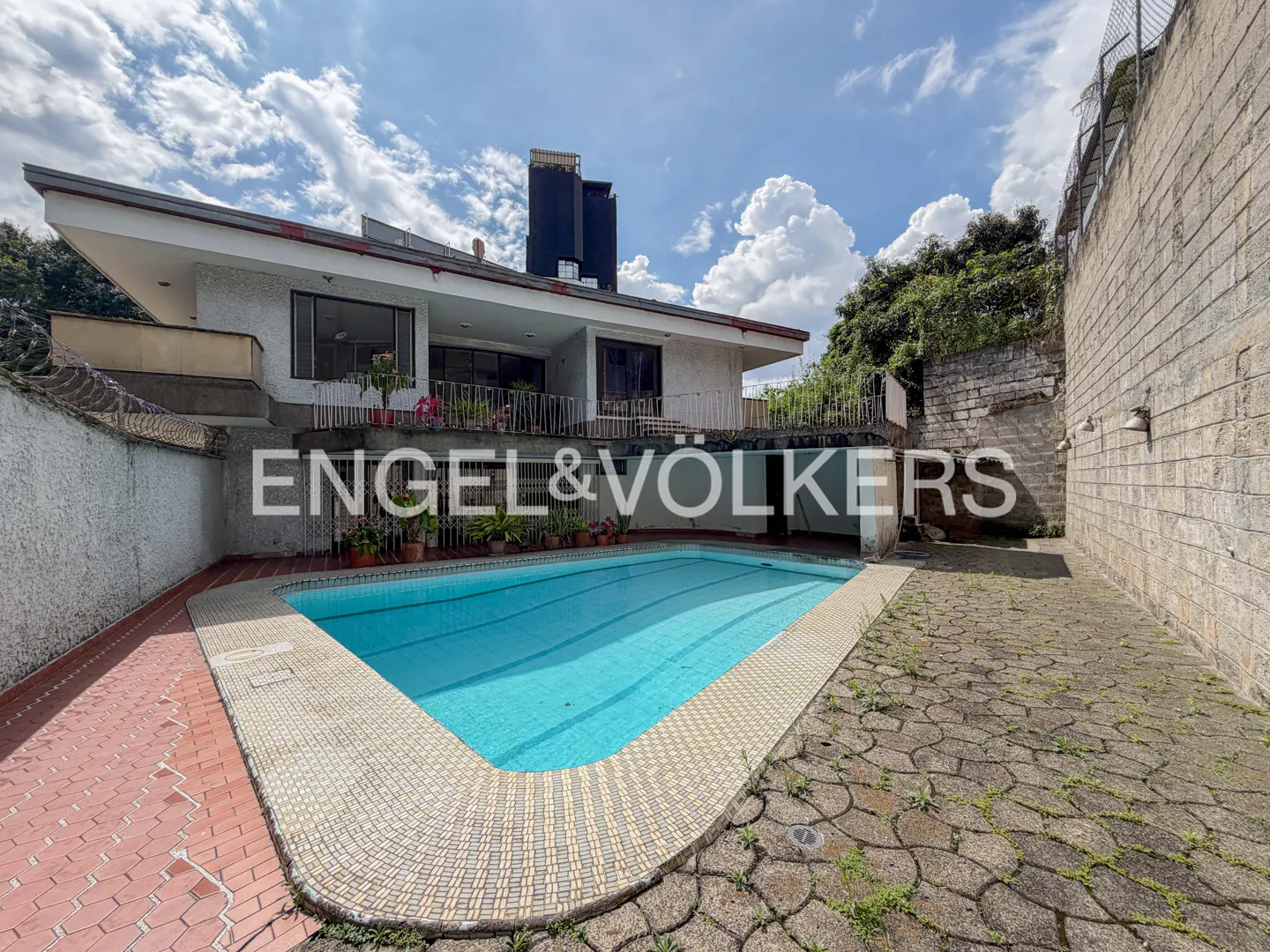 A backyard view of a two-story white house with a blue swimming pool and stone patio. The Engel & Völkers logo is superimposed on the image.