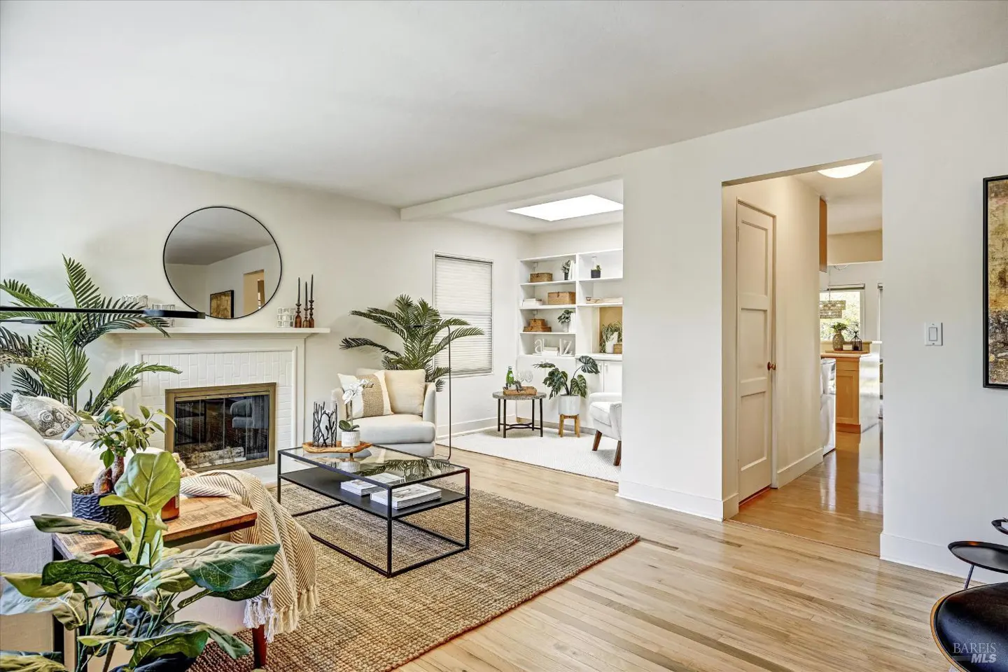 Bright living room with hardwood floors, white walls, fireplace, round mirror, plants, and a black metal coffee table.