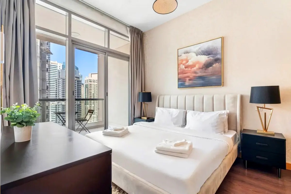 Bedroom with white linens, beige headboard, and balcony overlooking city skyline. Artwork hangs above the bed.