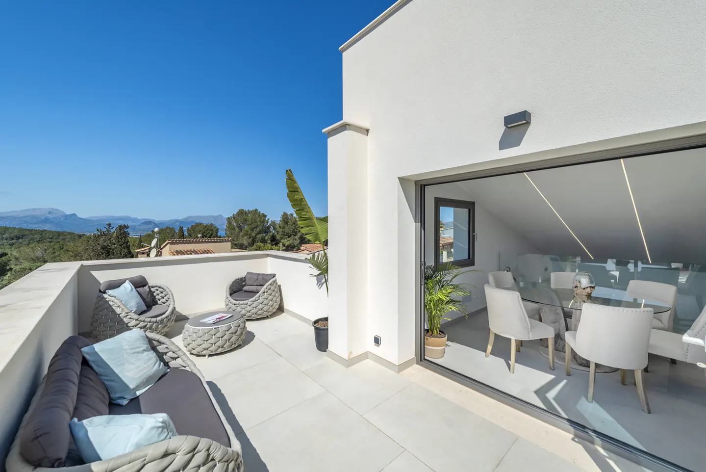 Outdoor patio with gray wicker furniture, blue pillows, and a view of mountains under a clear blue sky. Sliding glass doors lead to an indoor dining area.