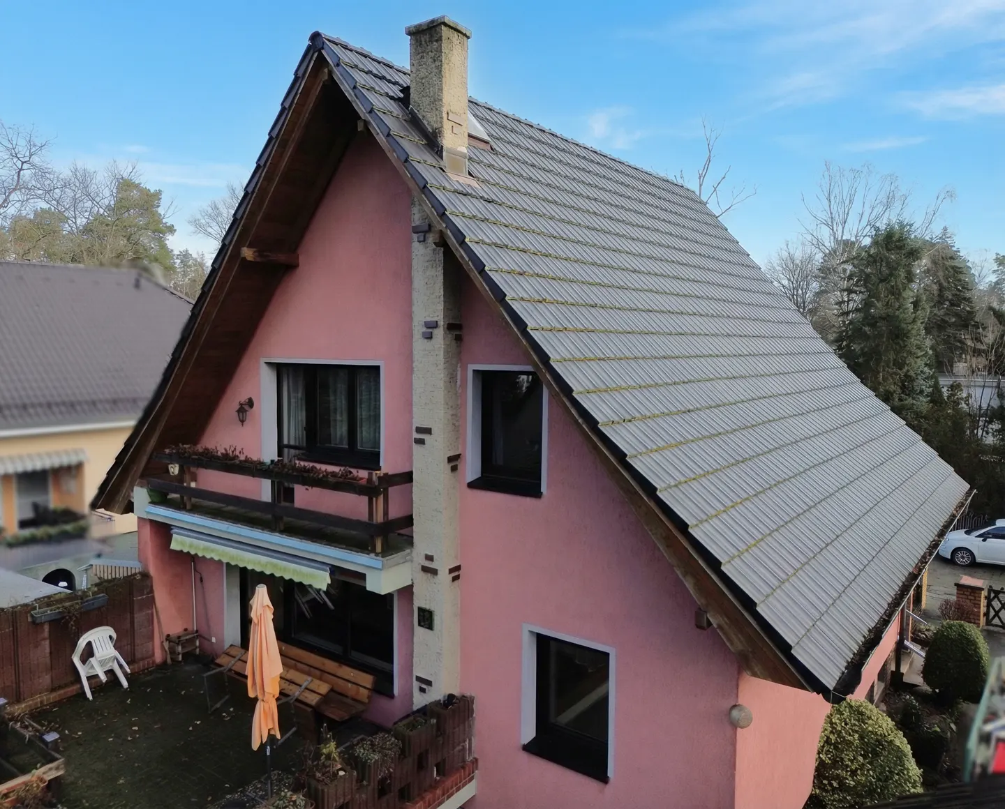 Two-story pink house with a gray tiled roof, chimney, and balcony. A patio with furniture is visible in the yard.