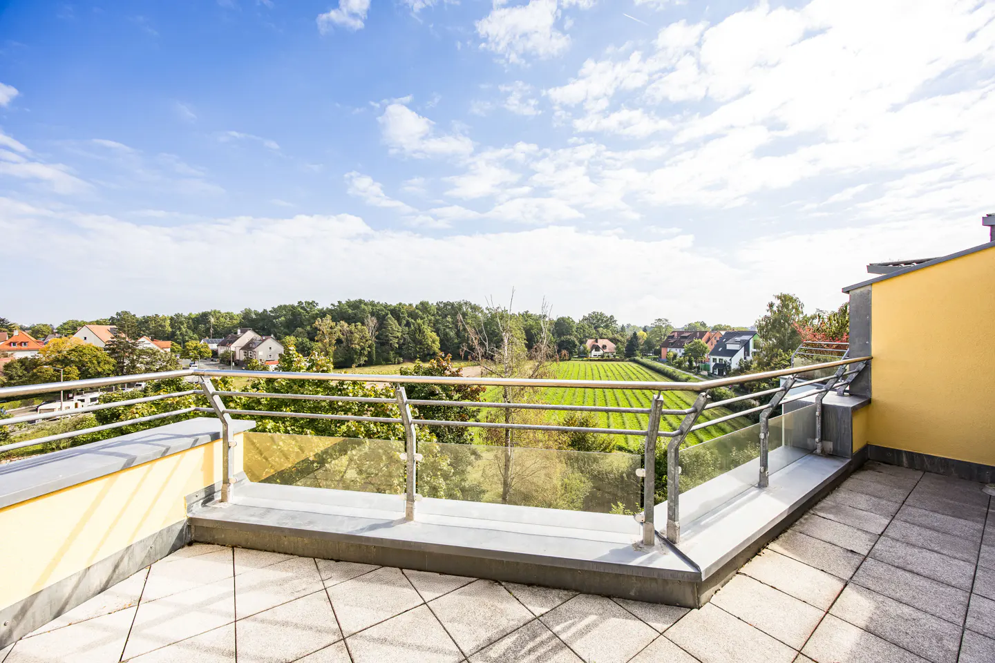 Balcony view of a green field and houses under a blue sky. The balcony has a gray tiled floor and a glass and metal railing.