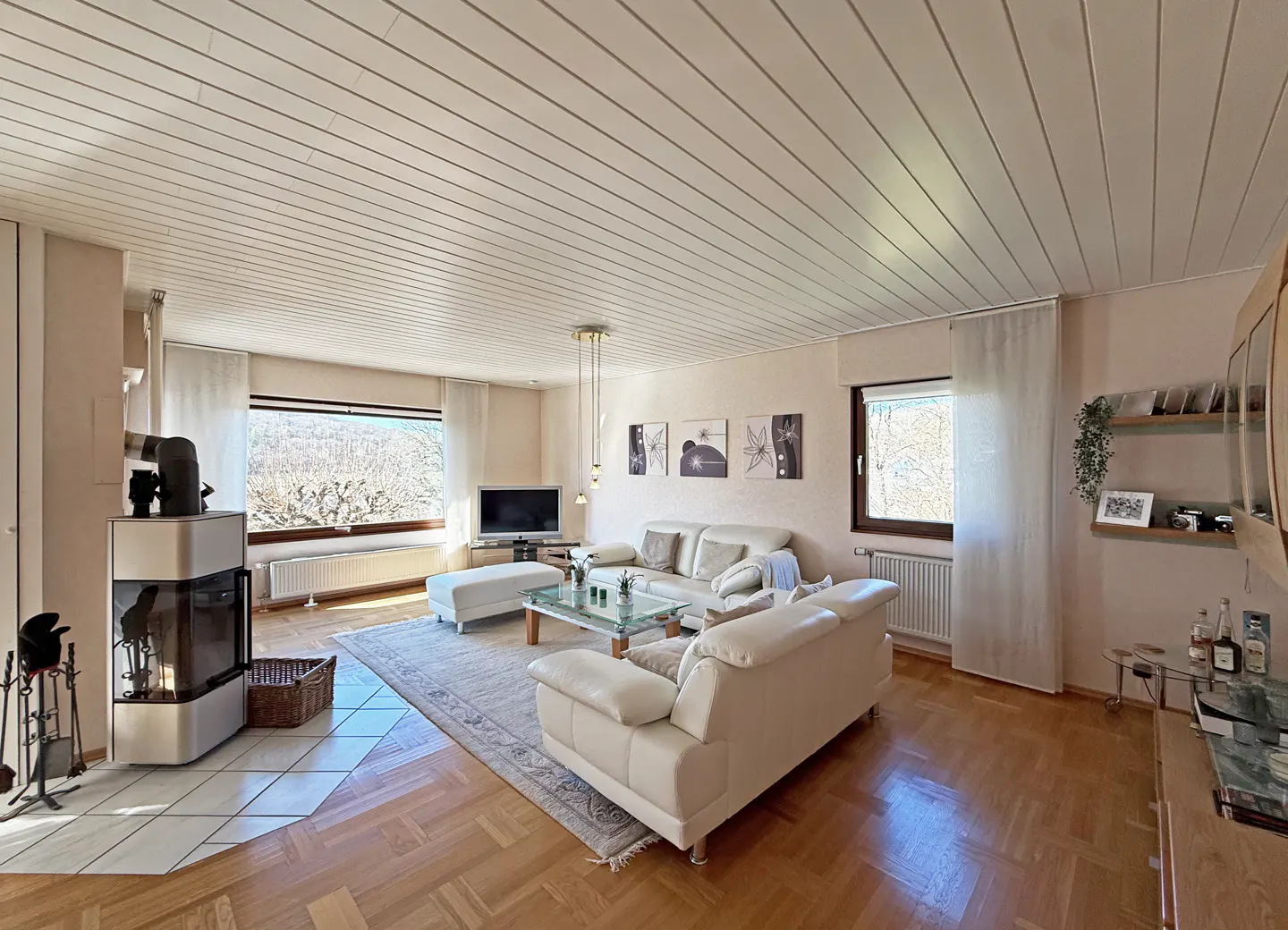 Bright living room with white sofas, glass table, and fireplace. Large windows offer natural light. Wood floors and ceiling add warmth.