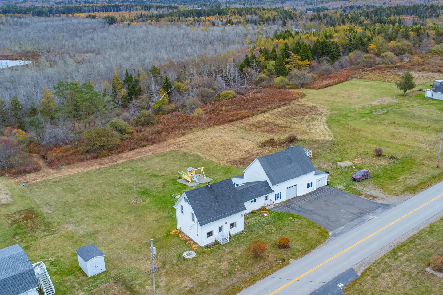 Aerial view of a white house with a gray roof, a playground, and a forest in the background.