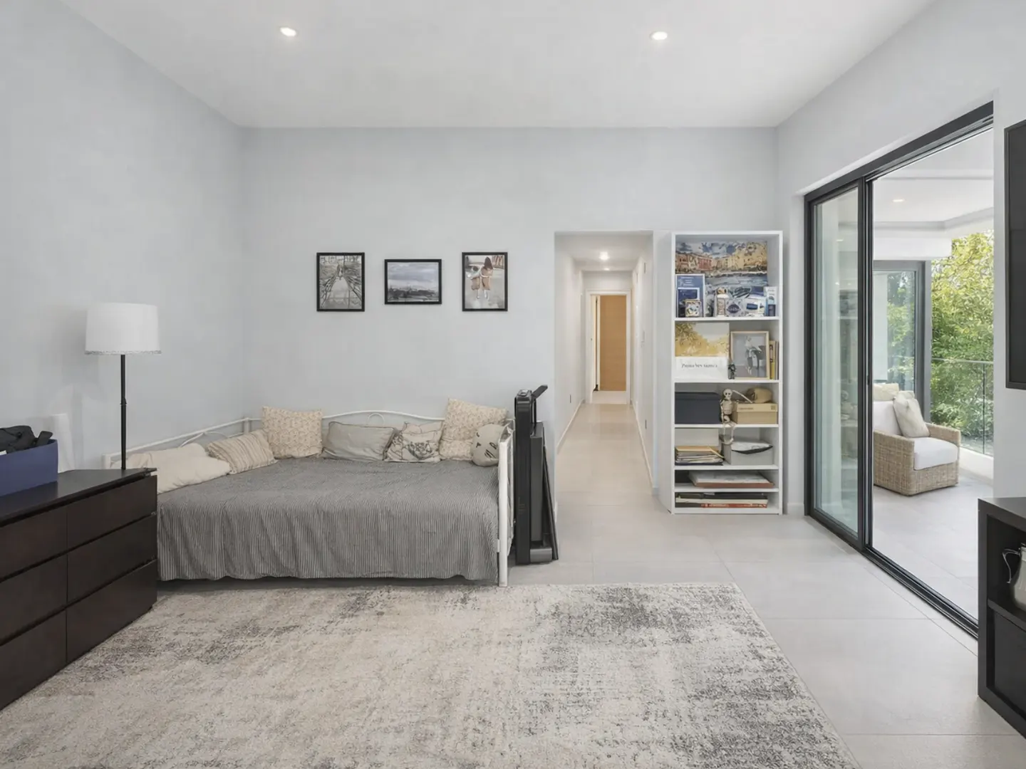 A bright room with a daybed, dresser, and bookshelf. Sliding glass doors lead to a patio with a wicker chair.