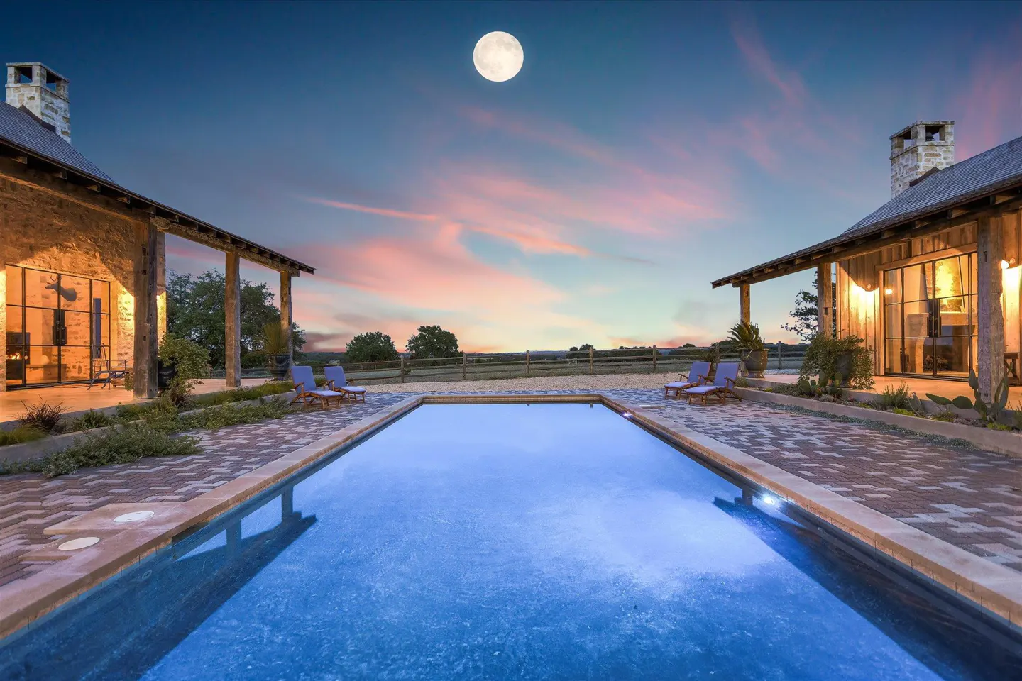 Twilight view of a rectangular pool between two stone buildings, with lounge chairs. A full moon is visible in the sky.