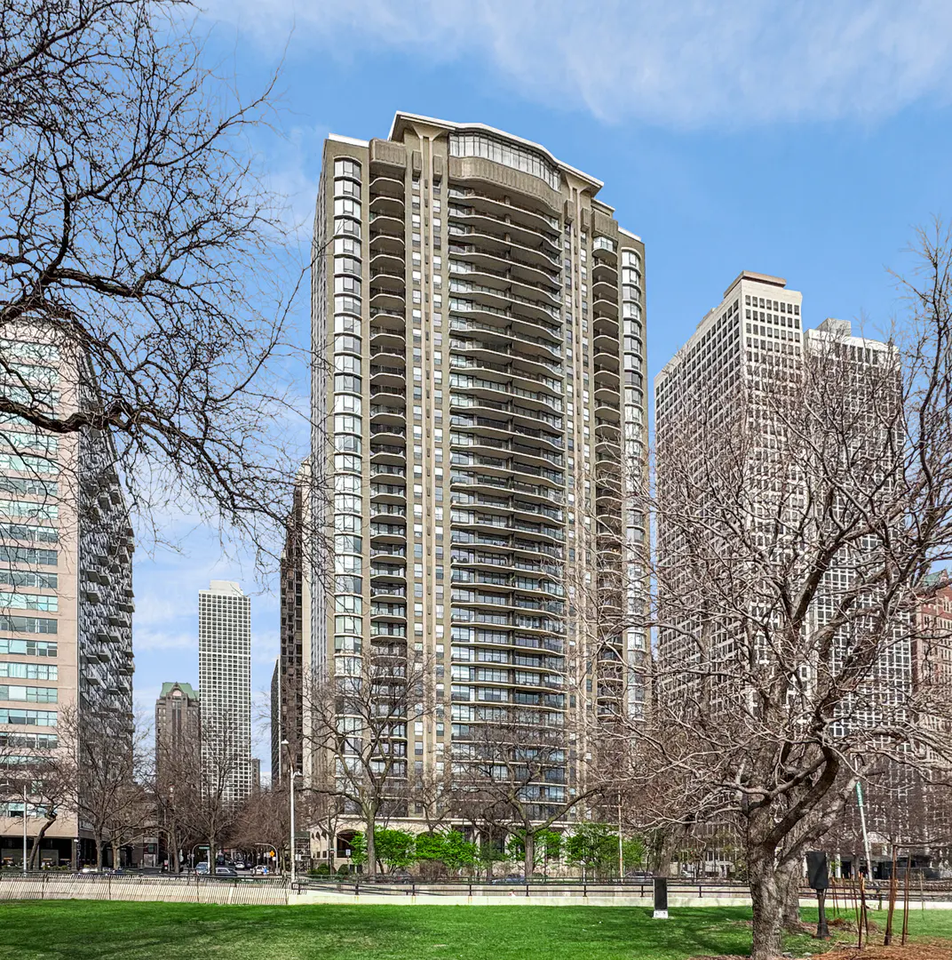 Exterior view of a tall, beige condo building with balconies, set against a blue sky and other buildings. Trees frame the view.