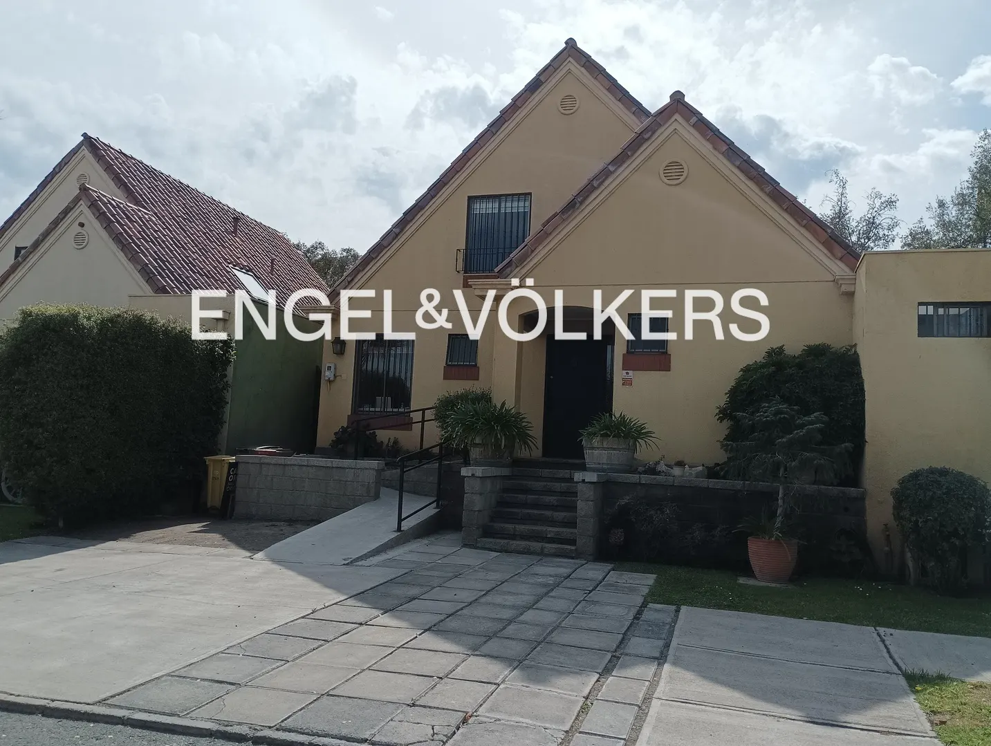 Exterior of a tan house with a red tile roof and black door. A stone walkway leads to the entrance. Bushes and trees surround the house.