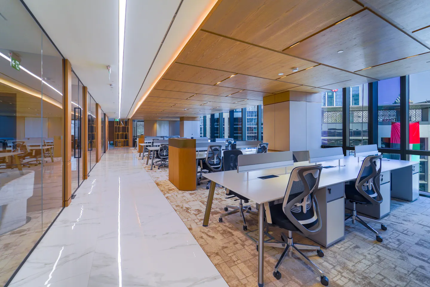 Bright, modern office space with white desks, gray chairs, and wood-paneled ceilings. Large windows offer a city view.