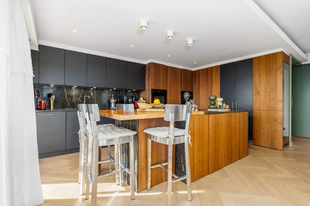 A modern kitchen with gray cabinets, wood island with metal stools, and marble backsplash.