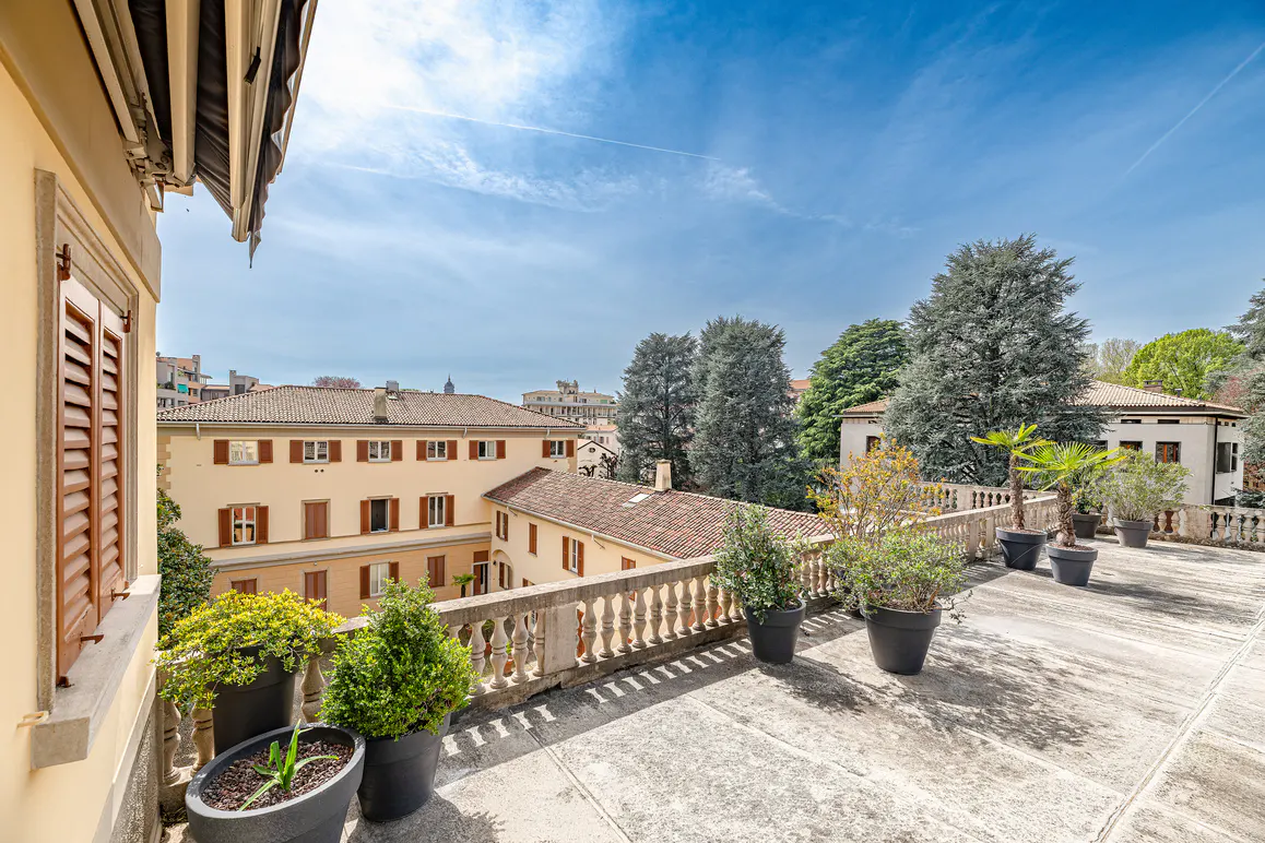 View from a balcony with potted plants, a stone balustrade, and buildings in the background under a blue sky.