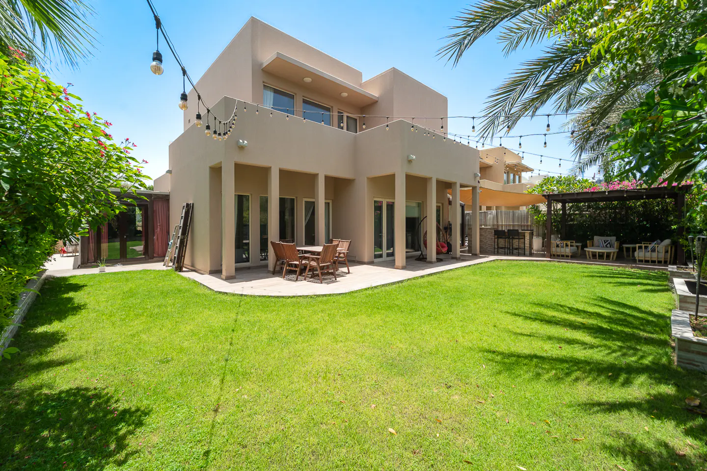 Backyard view of a two-story beige house with a green lawn, patio furniture, string lights, and lush greenery.