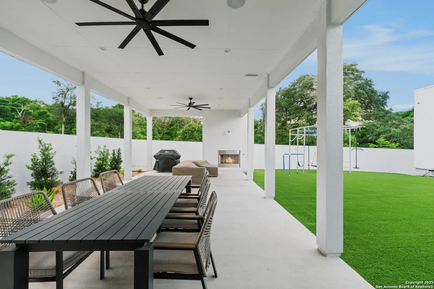 Covered patio with a long table, chairs, grill, and outdoor fireplace. Green lawn and trees in the background.