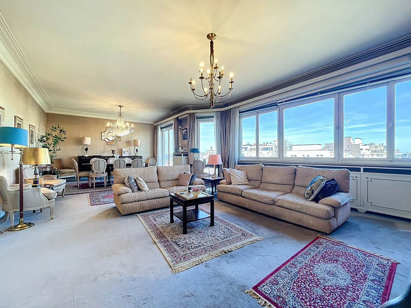 A spacious living room with beige sofas, patterned rugs, and large windows overlooking a cityscape. A chandelier hangs from the ceiling.