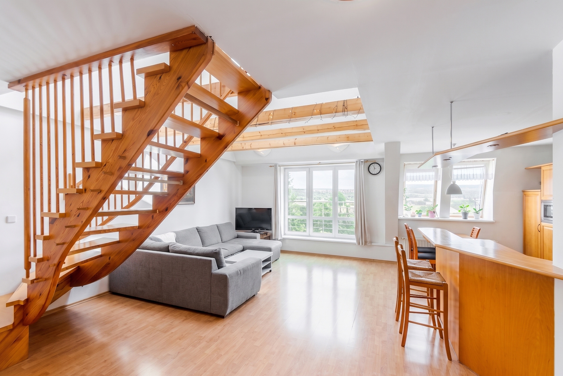 Open-plan living area with a wooden staircase, grey sectional sofa, and a kitchen island with bar stools on wood floors.