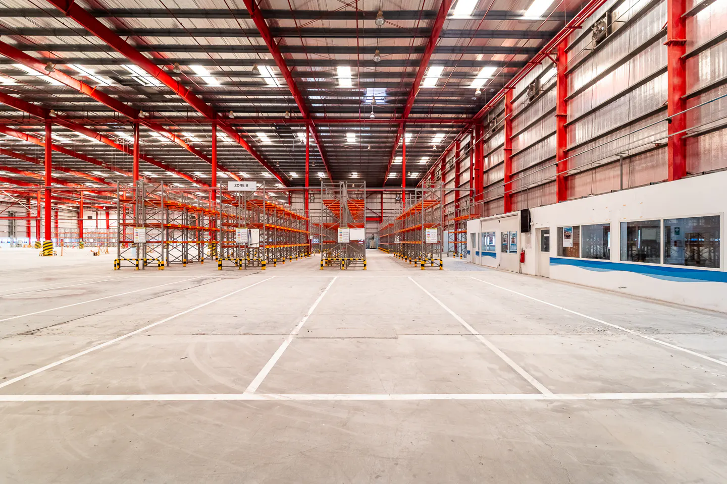 A large, empty warehouse with red support beams and rows of metal shelving. White lines mark parking spaces on the concrete floor.