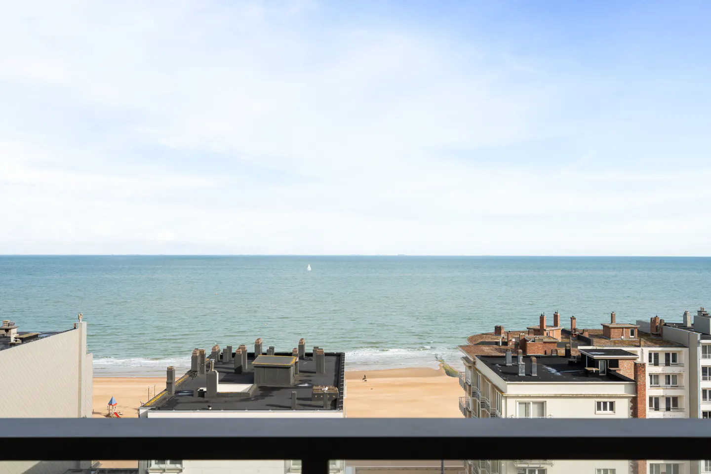 Elevated view from a balcony overlooking a sandy beach, the ocean, and nearby apartment buildings under a bright sky.