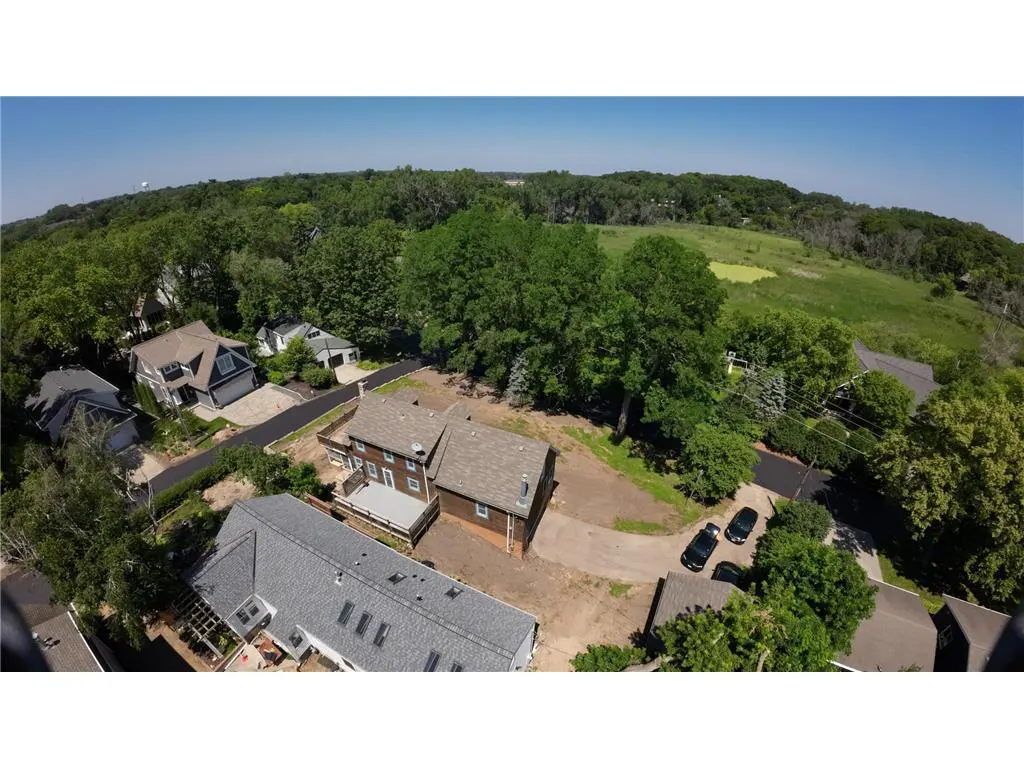 Aerial view of a brown two-story house with a gray roof, surrounded by green trees and a grassy field. Two black cars are parked in the driveway.
