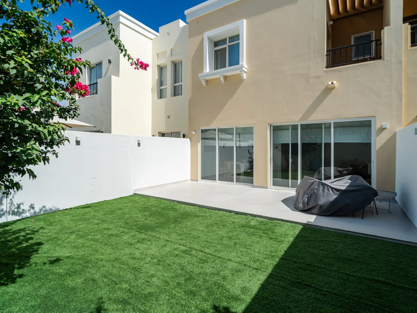 Backyard view of a two-story beige house with a green lawn, white wall, and sliding glass doors. A covered outdoor furniture set sits on the patio.