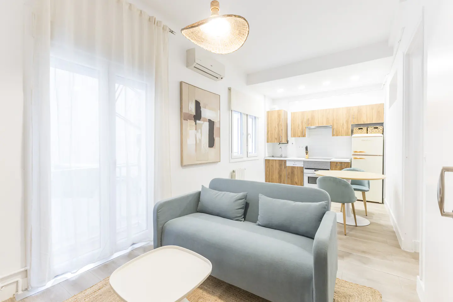Bright apartment interior with a blue sofa, white coffee table, and open kitchen with wood cabinets and a retro fridge.