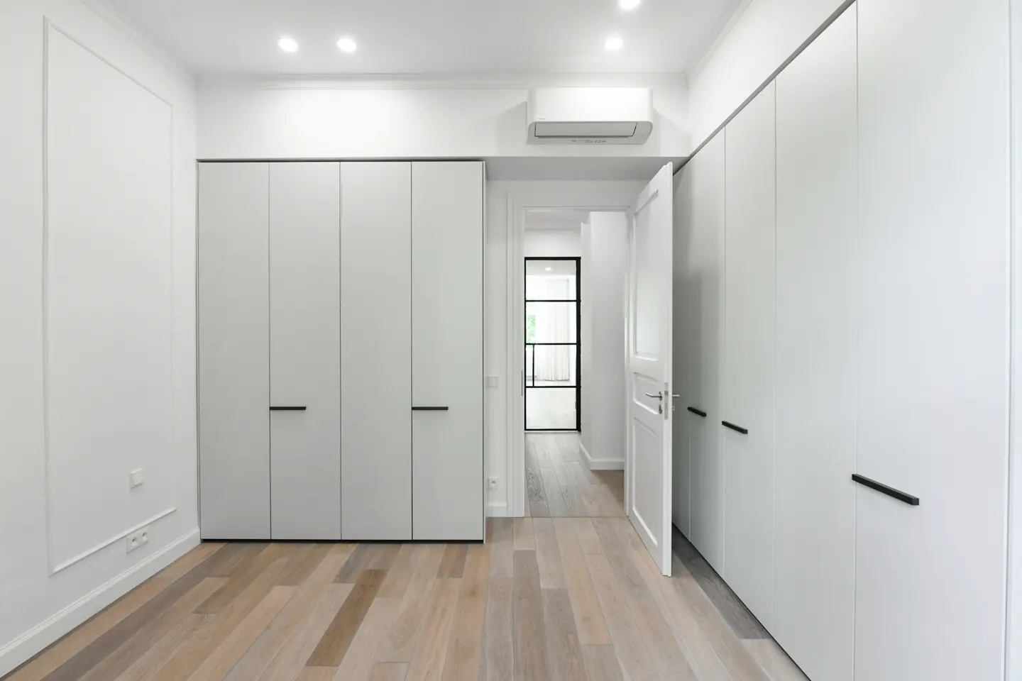 Bright, modern room with light wood floors, white walls, and tall white wardrobes with black handles. A doorway leads to another room.