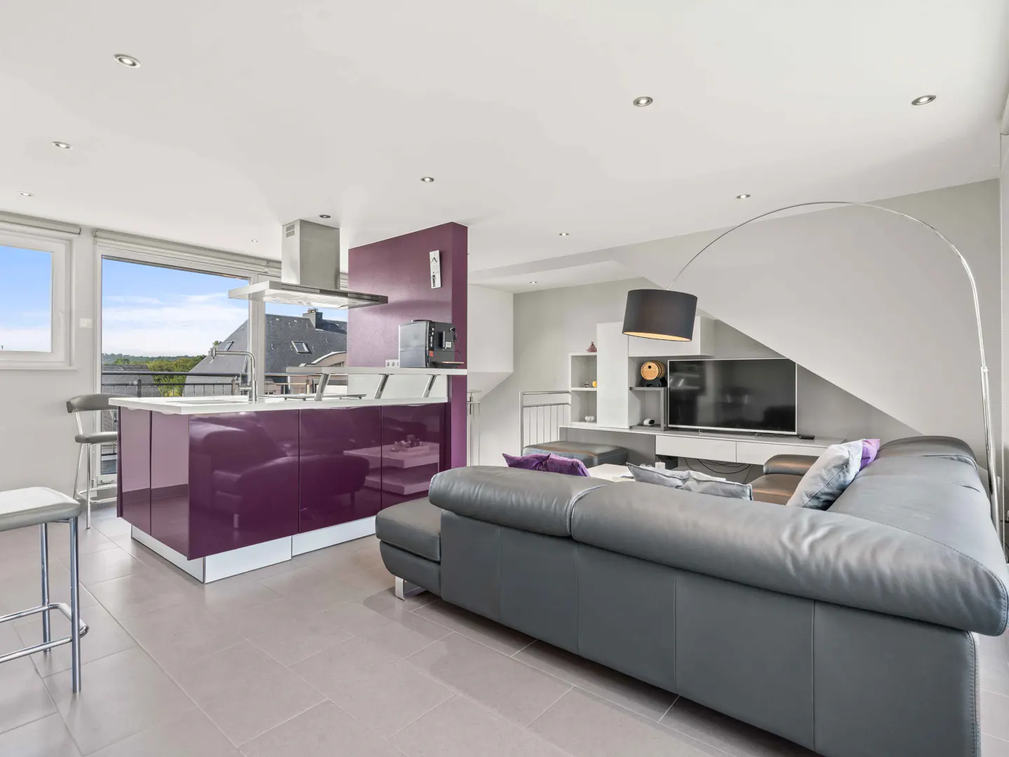 Open-concept living space with a purple kitchen island, gray leather sofa, and a large window with a view.