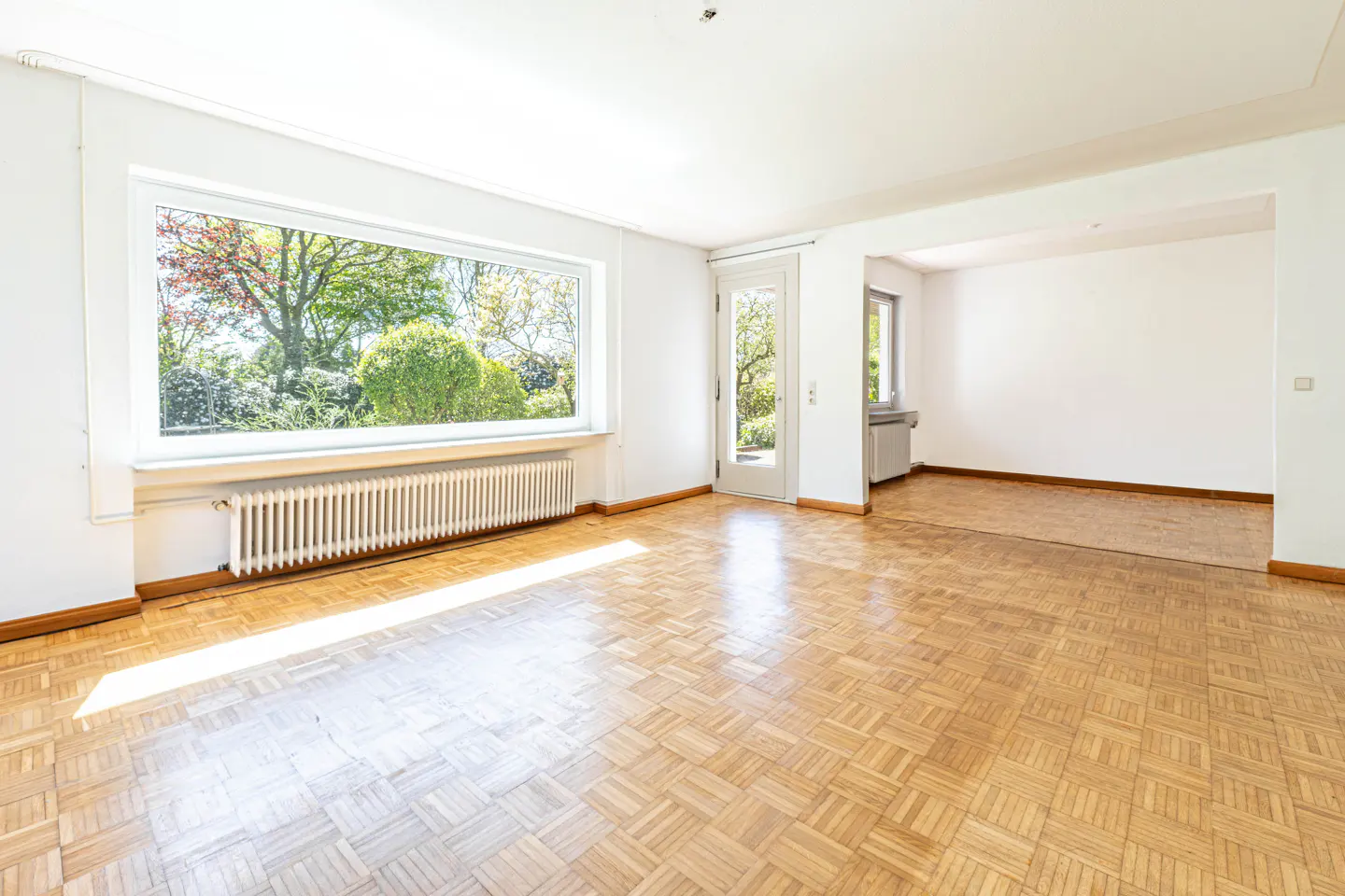 Bright, empty room with parquet floors, white walls, large window overlooking green garden, and a white door.
