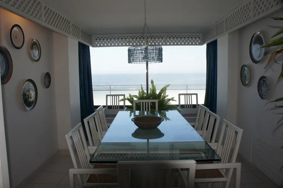 Dining room with a glass table, white chairs, and ocean view. Plates decorate the white walls. A fern sits in the center of the table.