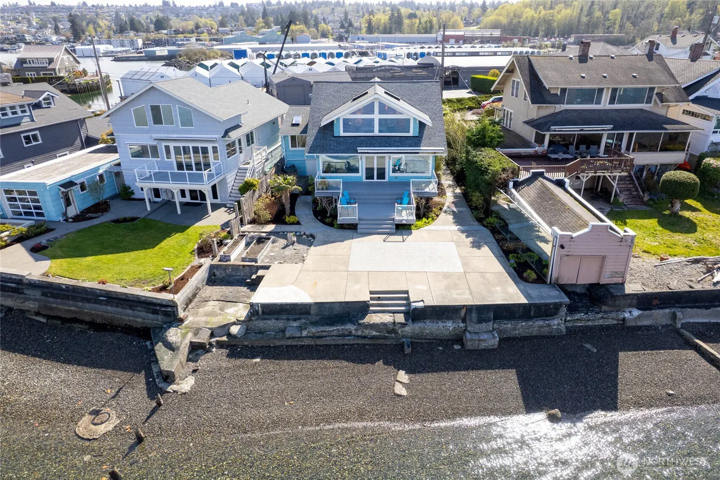 Aerial view of a light blue waterfront house with a gray roof, a concrete patio, and a beach in front.