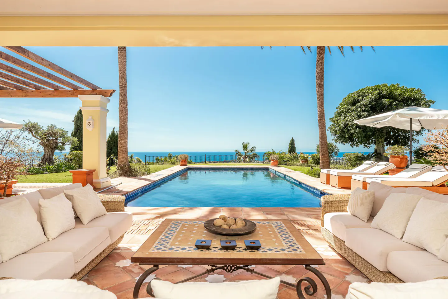 Outdoor patio with white sofas, a patterned table, and a pool overlooking the ocean on a sunny day.