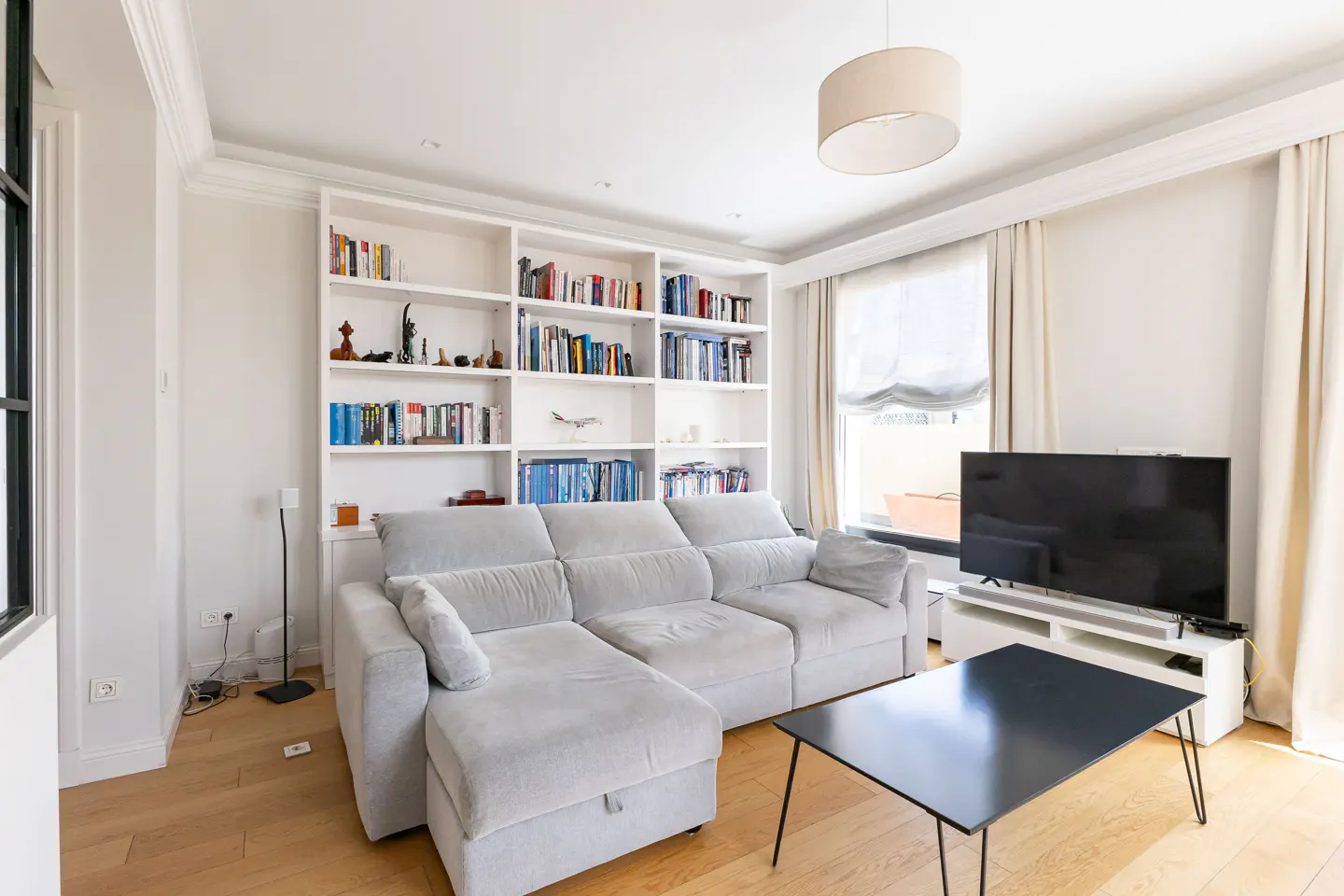 Bright living room with a light gray sectional sofa, a black coffee table, a large bookshelf, and a flat-screen TV.