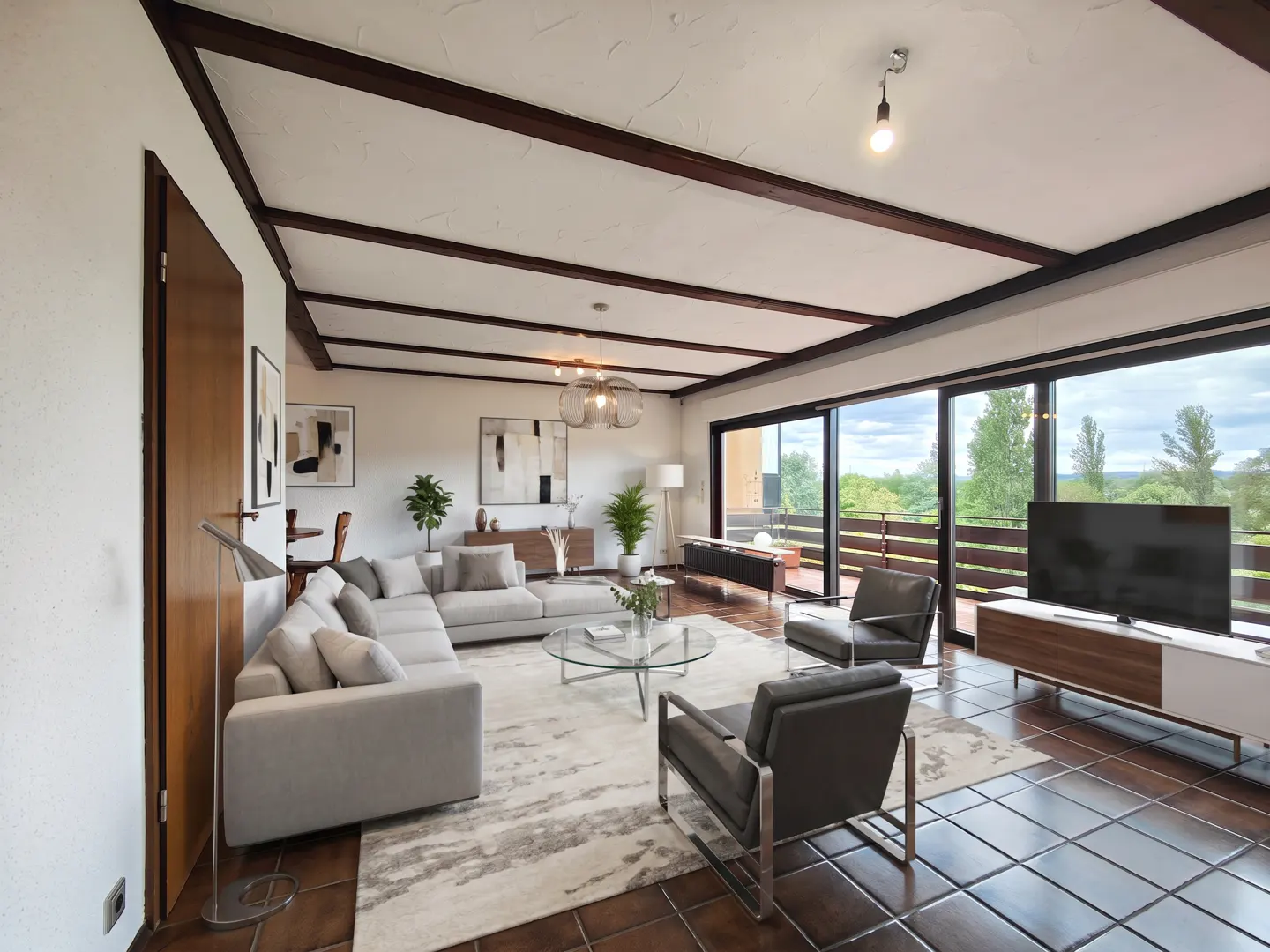 Bright living room with a gray sectional sofa, glass table, and dark wood ceiling beams. Large windows overlook a green landscape.