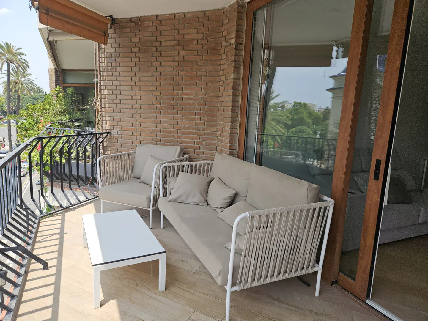 Balcony with beige outdoor sofa, armchair, and white coffee table against a brick wall, overlooking a street with palm trees.