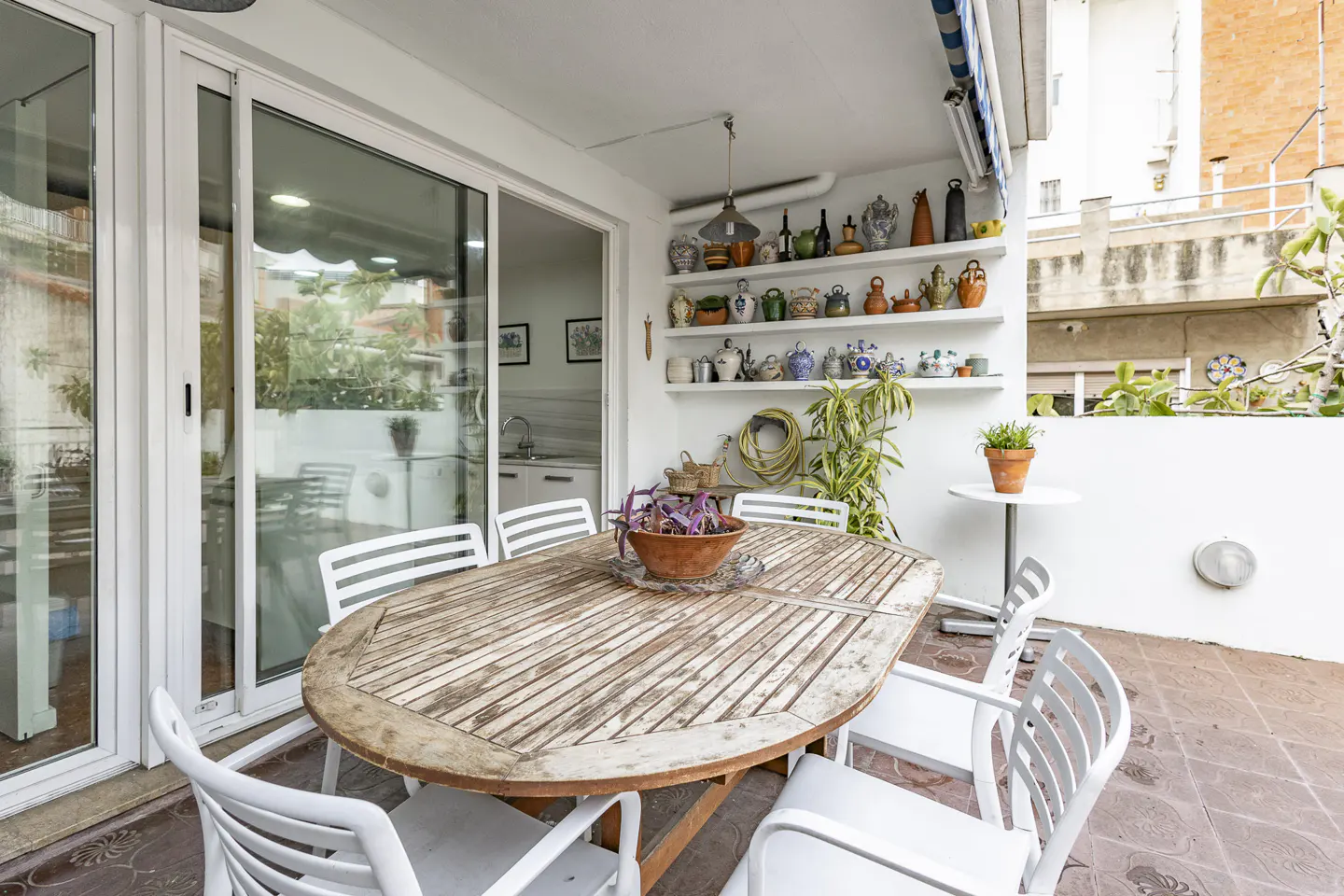 Outdoor patio with a weathered wood table, white chairs, and shelves displaying decorative items against a white wall.