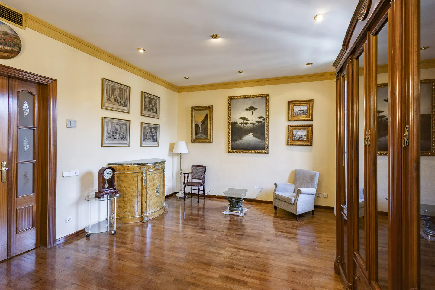 A formal living room with hardwood floors, cream walls, and framed art. A wooden armoire stands to the right.