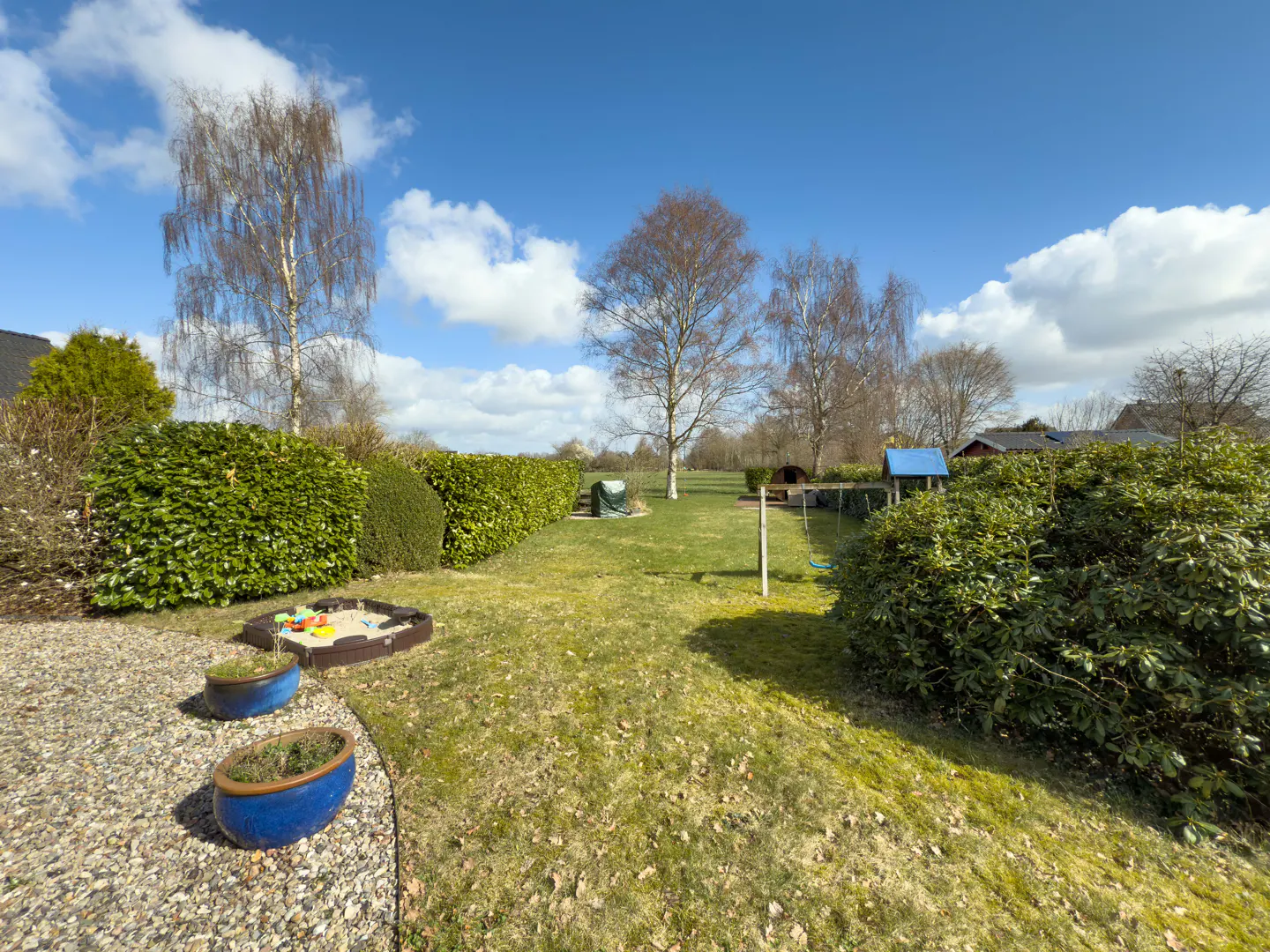 A sunny backyard with green grass, trees, and a blue sky with white clouds. A sandbox and blue planters are in the foreground.