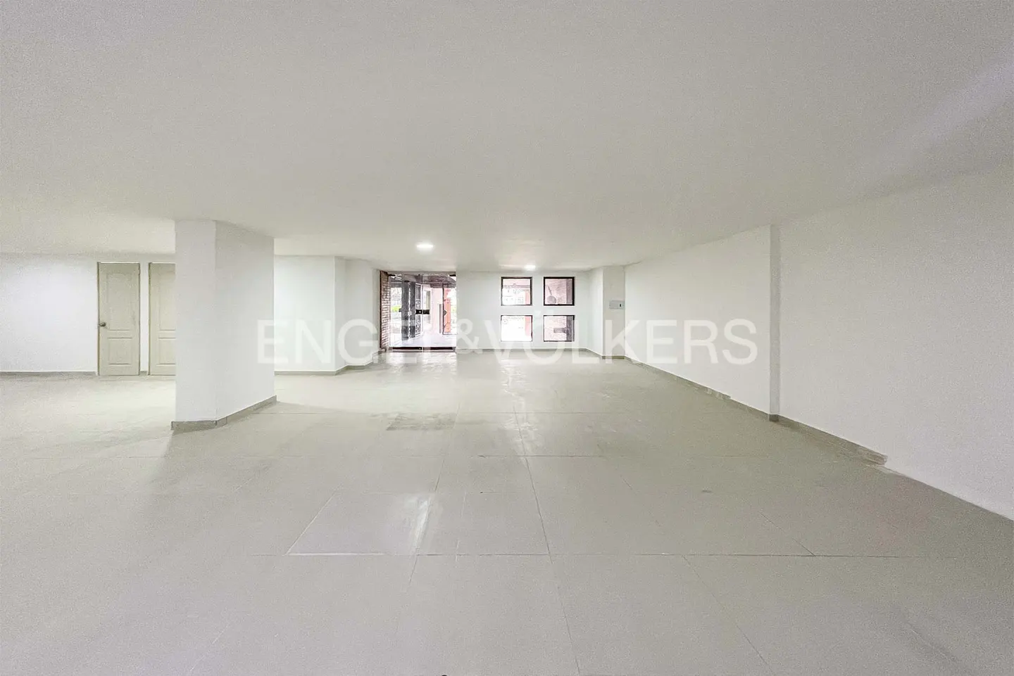 A bright, empty room with white walls and a light gray tiled floor. A doorway is visible in the back.