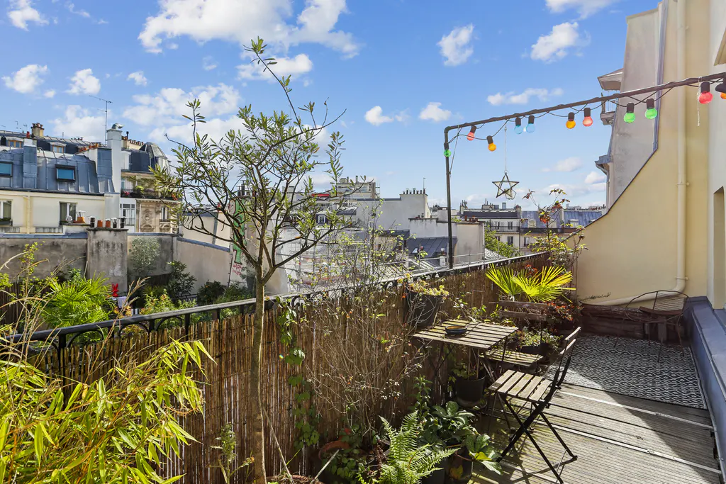 A Parisian balcony with a small table, chairs, and plants, overlooking city rooftops under a blue sky with clouds. String lights add a festive touch.