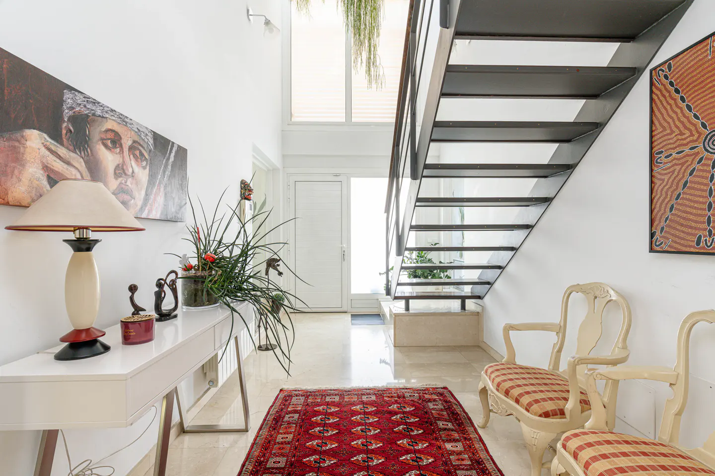 Bright foyer with white walls, marble floors, and a red patterned rug. A modern staircase, console table with lamp, and artwork add character.