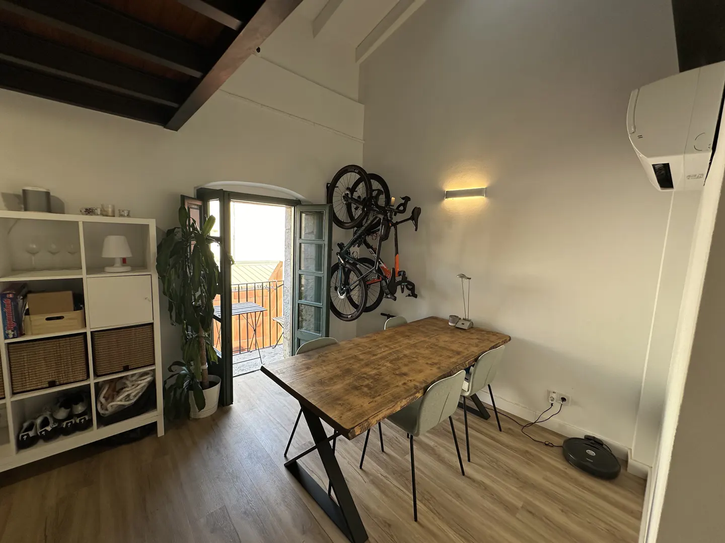 A bright room with a wooden table, four chairs, and two bikes mounted on the wall. A balcony is visible through an open door.