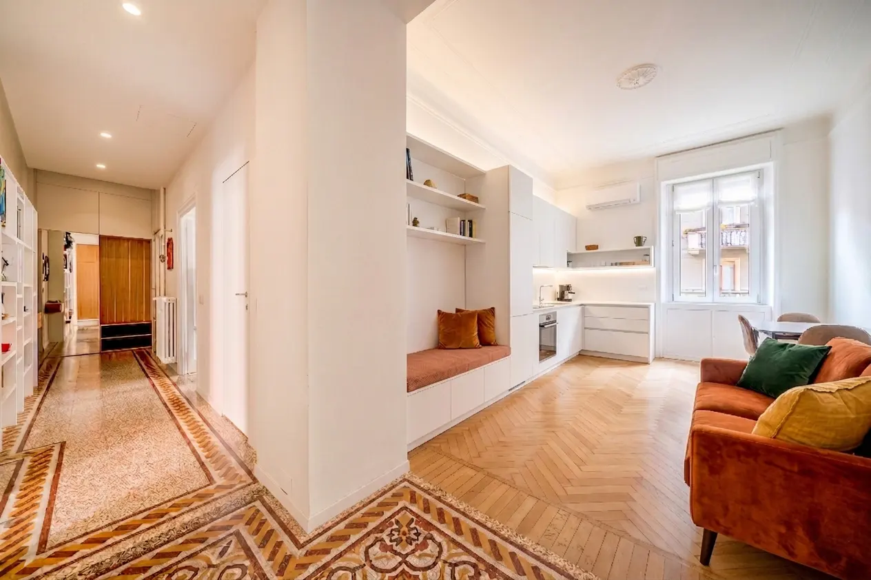 Modern apartment interior with a hallway featuring patterned tile floors, a white kitchen, and a rust-colored sofa.