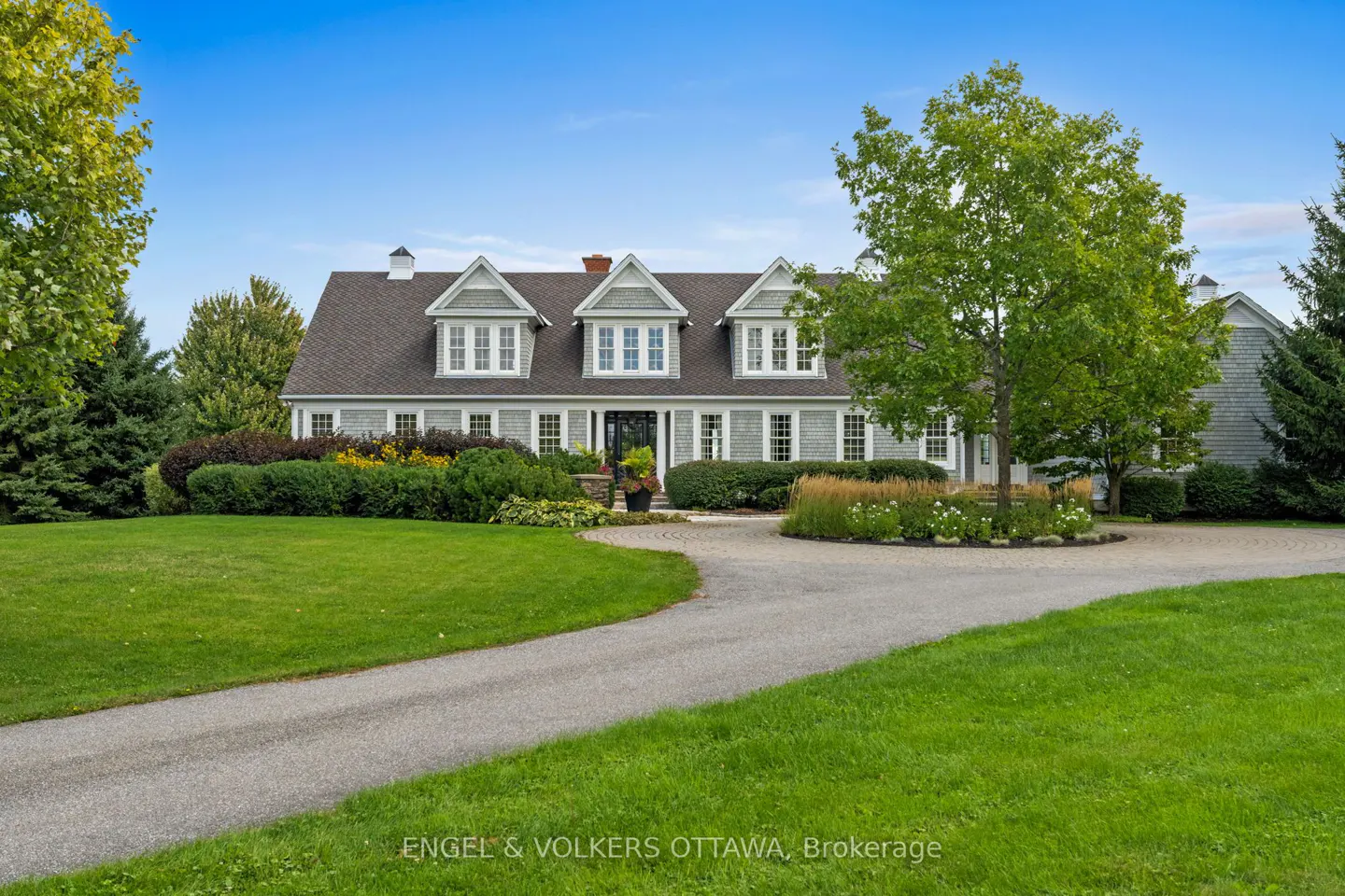 Exterior view of a gray house with a dark roof, green lawn, and circular driveway on a sunny day.