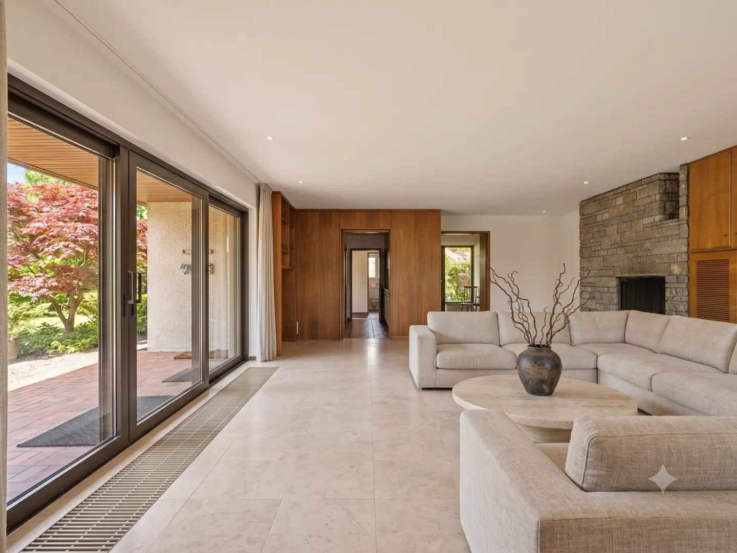 Modern living room with beige sectional sofa, stone fireplace, wood paneling, and sliding glass doors to a garden.