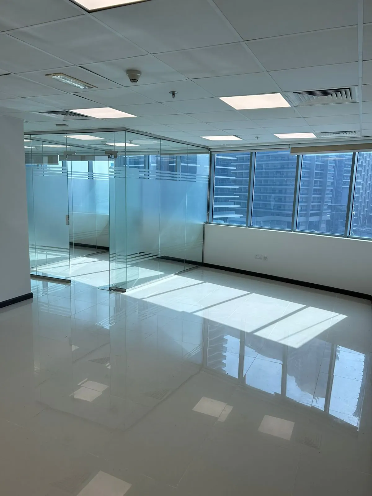 Empty office space with glass-walled rooms, a shiny floor, and large windows showing a city skyline.