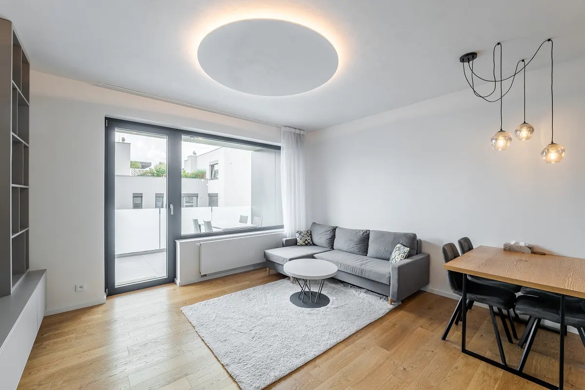 Bright living room with wood floors, gray sofa, white rug, and dining table. Large window with balcony view. Modern lighting fixtures.