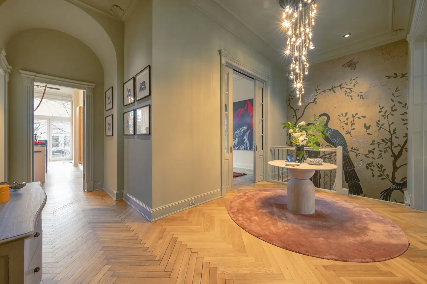 Hallway with herringbone wood floors, pale green walls, and a chandelier. A round table sits on a pink rug, with a mural of a peacock behind it.
