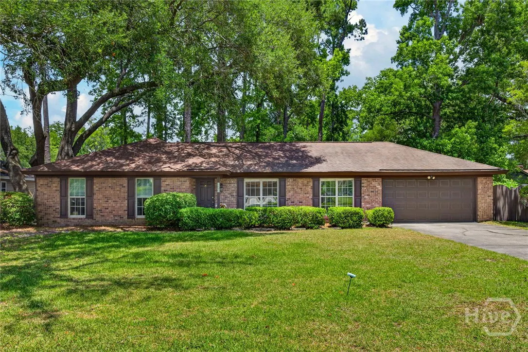 Single-story brick house with brown roof, shutters, and garage door. Green lawn and trimmed bushes in front. Trees in background.