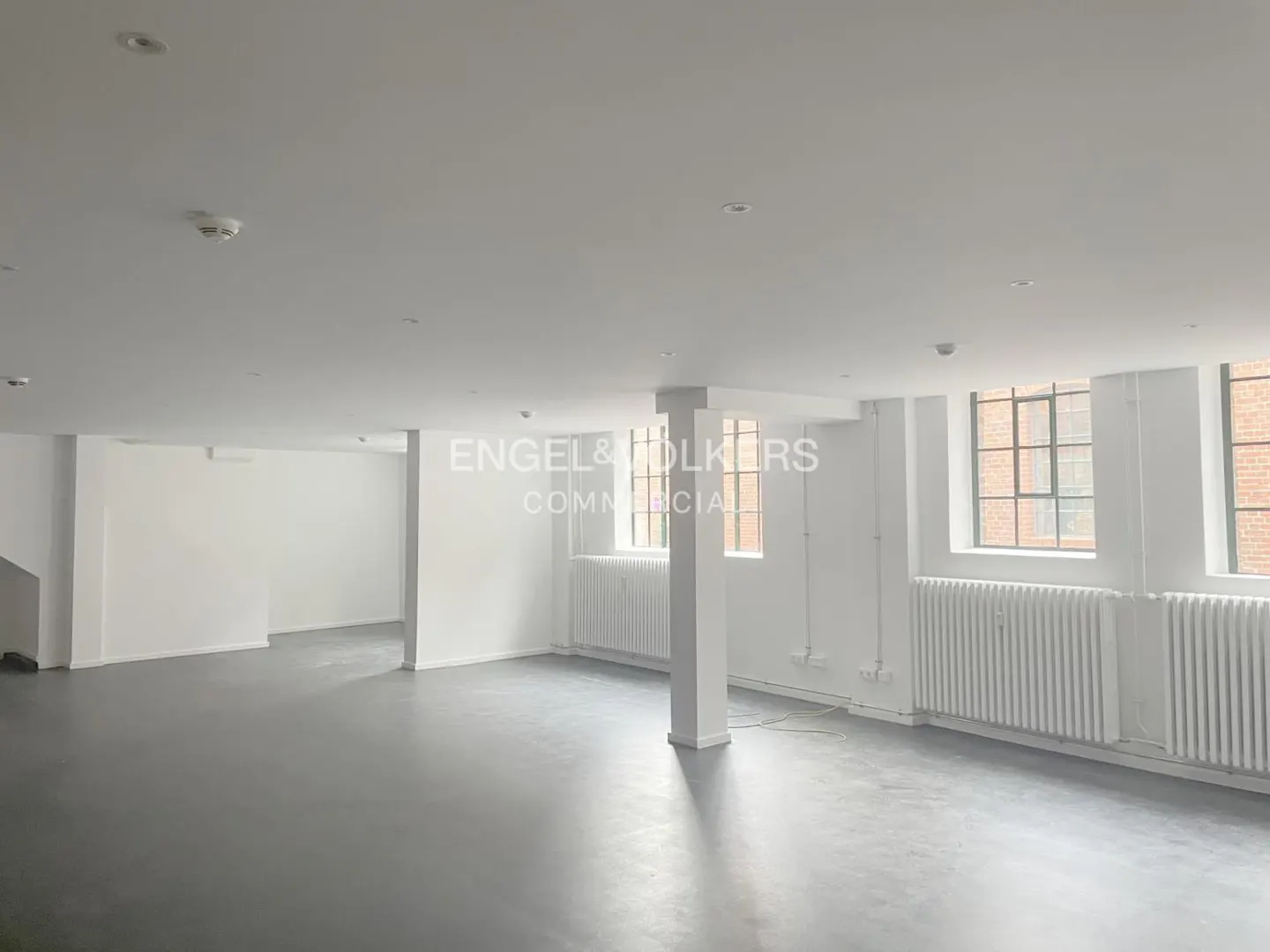 Bright, empty commercial space with gray floors, white walls, and large windows. Radiators line the walls below the windows. Pillar in the center.